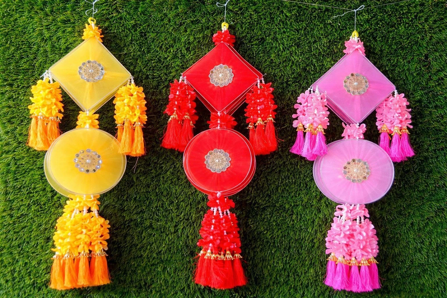 Diamond and round satin Diwali toran hanging with sequin medallion centres and fabric flower tassels, shown in yellow, red and pink